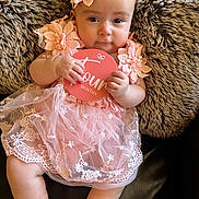 Ariana is registered to the contest to win money with this photo: baby, infant, pink_dress, headband, flower, sign, months, fur_cushion, leather_couch, holding, portrait, sitting, cute, child, person, indoors, soft_texture, smiling, young, closeup