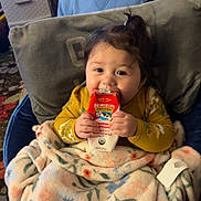 Neala is registered to the contest to win money with this photo: baby, child, blanket, milk_carton, organic_milk, person, indoor, cozy, floral_pattern, clothing, seated, portrait, face, hand, looking, young_child, toddler, table, furniture, warm