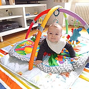 Kylian participe au concours pour gagner de l'argent avec cette photo : baby, play_mat, activity_gym, indoor, child, smiling, colorful, toy, floor, window, curtain, radiator, television, furniture, shelf, home, person, bib, striped_clothing, light