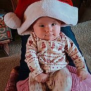 Estella is registered to the contest to win money with this photo: baby, child, santa_hat, pajamas, indoor, carpet, person, lap, smile, cute, festive, patterned_clothing, furniture, cozy, holiday, red_hat, white_hat, legs, candid, portrait