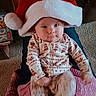 baby, child, santa_hat, pajamas, indoor, carpet, person, lap, smile, cute, festive, patterned_clothing, furniture, cozy, holiday, red_hat, white_hat, legs, candid, portrait