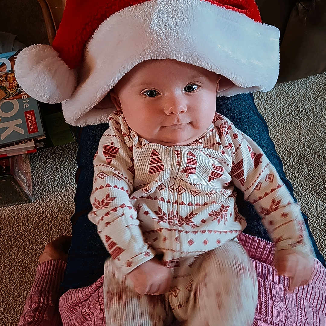 Estella is registered to the contest to win money with this photo: baby, child, santa_hat, pajamas, indoor, carpet, person, lap, smile, cute, festive, patterned_clothing, furniture, cozy, holiday, red_hat, white_hat, legs, candid, portrait