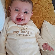Myriam a rejoint le concours — aidez-le/la à gagner de superbes lots ! baby, smiling, onesie, pillow, yellow_pillow, white_clothing, infant, happy, couch, face, child, lying_down, indoors, cute, expression, hand, soft_texture, cozy, young_child, portrait
