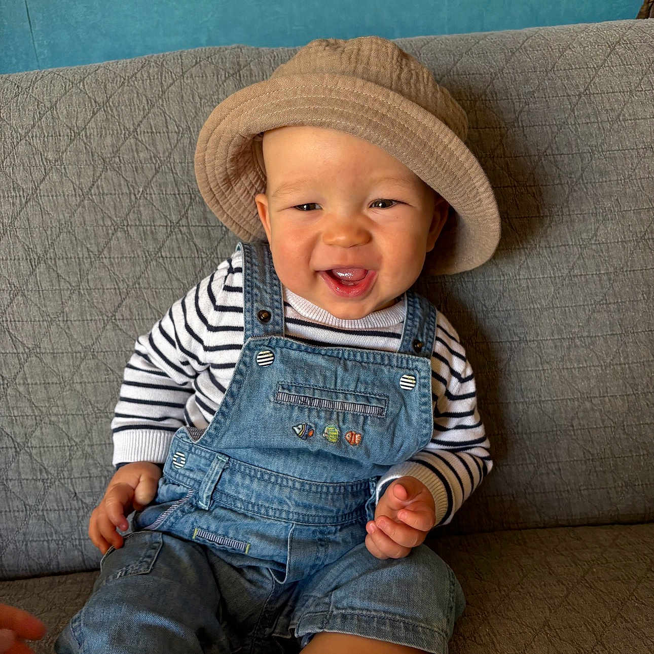Maël Roux participe au concours pour gagner de l'argent avec cette photo : toddler, child, smiling, hat, bucket_hat, denim_overalls, striped_shirt, sitting, couch, indoor, happy, portrait, baby, face, clothing, furniture, skin, person, casual, cute