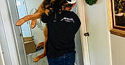 Kane joined the competition — help win amazing prizes! person, dog, hallway, cap, black_shirt, metallica, carrying, walking, wreath, framed_painting, door, wood_floor, indoor, relaxed_dog, casual_clothing, back_view, large_dog, white_cap, decor, home