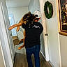person, dog, hallway, cap, black_shirt, metallica, carrying, walking, wreath, framed_painting, door, wood_floor, indoor, relaxed_dog, casual_clothing, back_view, large_dog, white_cap, decor, home