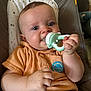 baby, child, teething_toy, orange_shirt, dinosaur_badge, cushioned_seat, happy, smiling, portrait, indoor, hands, face, cute, infant, chewing, blue_eyes, skin, short_hair, clothing, baby_gadget