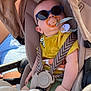 baby, sunglasses, pacifier, stroller, child, yellow_shirt, green_shorts, harness, seat, sunlight, outdoor, infant, cute, relaxed, fashion, shade, comfort, toy, person, summer