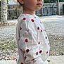 child, toddler, outdoor, tree, fence, apple_pattern, white_clothing, long_sleeve, pants, curly_hair, standing, side_profile, expression, nature, ground, concrete, leaves, daylight, casual, portrait