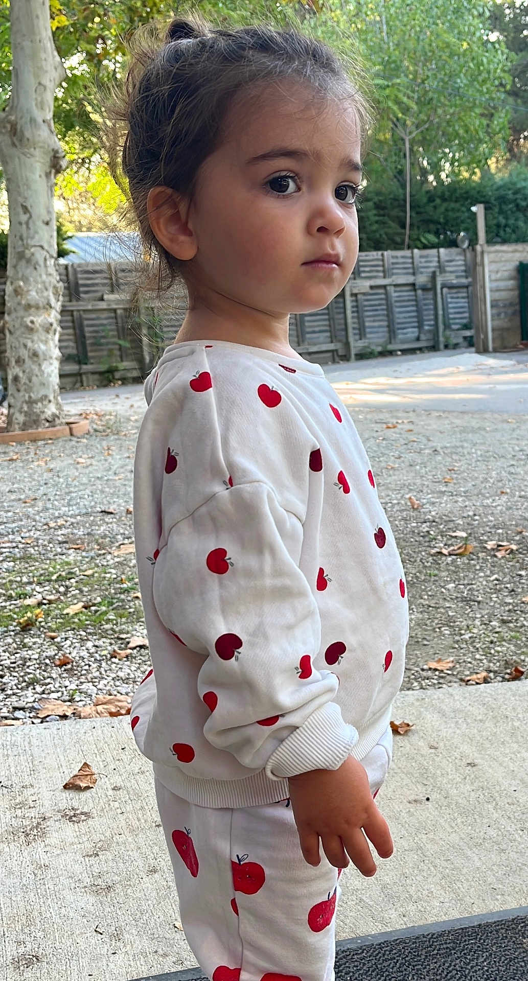Saona participe au concours pour gagner de l'argent avec cette photo : child, toddler, outdoor, tree, fence, apple_pattern, white_clothing, long_sleeve, pants, curly_hair, standing, side_profile, expression, nature, ground, concrete, leaves, daylight, casual, portrait