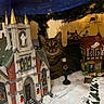 cat, tabby_cat, miniature_village, christmas_tree, holiday_decorations, snow, lamp_post, church, house, ornament, indoor, festive, lighting, curious, pet, animal, decoration, cozy, table, scene