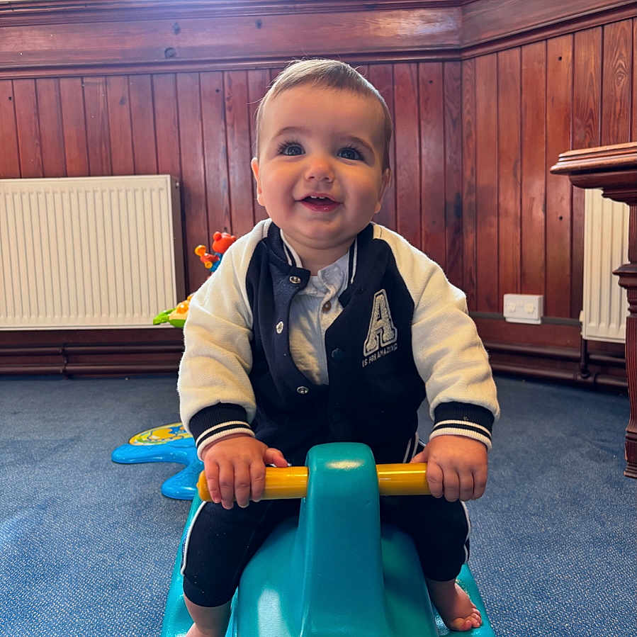 Oakley is registered to the contest to win money with this photo: baby, child, smiling, indoor, toy, rocking_toy, blue, wood_paneling, carpet, happy, cute, playful, clothing, jacket, hands, feet, furniture, room, person, portrait