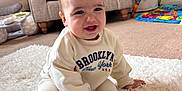 Oakley is registered to the contest to win money with this photo: baby, child, sitting, smiling, indoor, rug, cozy, sweatshirt, sock, toy, plush, furniture, carpet, living_room, cute, young_child, happy, face, person, home