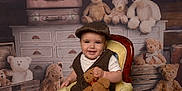 Oakley is registered to the contest to win money with this photo: baby, child, teddy_bear, chair, smile, vintage_clothing, hat, stuffed_animal, attic, suitcase, wooden_furniture, plush_toy, portrait, indoor, cute, happy, floor_rug, seated, person, cozy