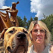 Toke is registered to the contest to win money with this photo: dog, person, selfie, outdoor, statue, blue_sky, clouds, sunlight, glasses, blonde_hair, tank_top, trees, stone_wall, portrait, closeup, daylight, nature, sculpture, casual, happy