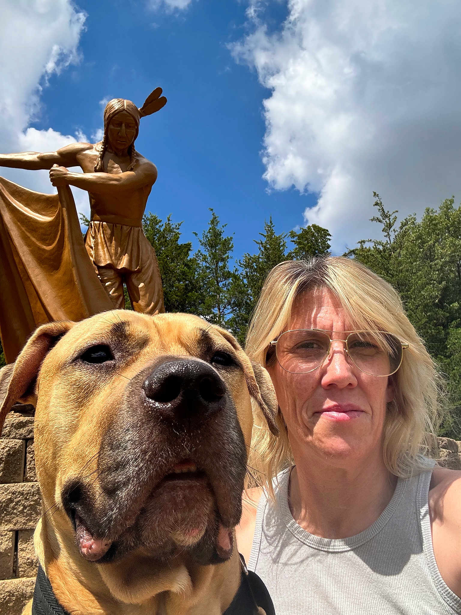 Toke is registered to the contest to win money with this photo: dog, person, selfie, outdoor, statue, blue_sky, clouds, sunlight, glasses, blonde_hair, tank_top, trees, stone_wall, portrait, closeup, daylight, nature, sculpture, casual, happy