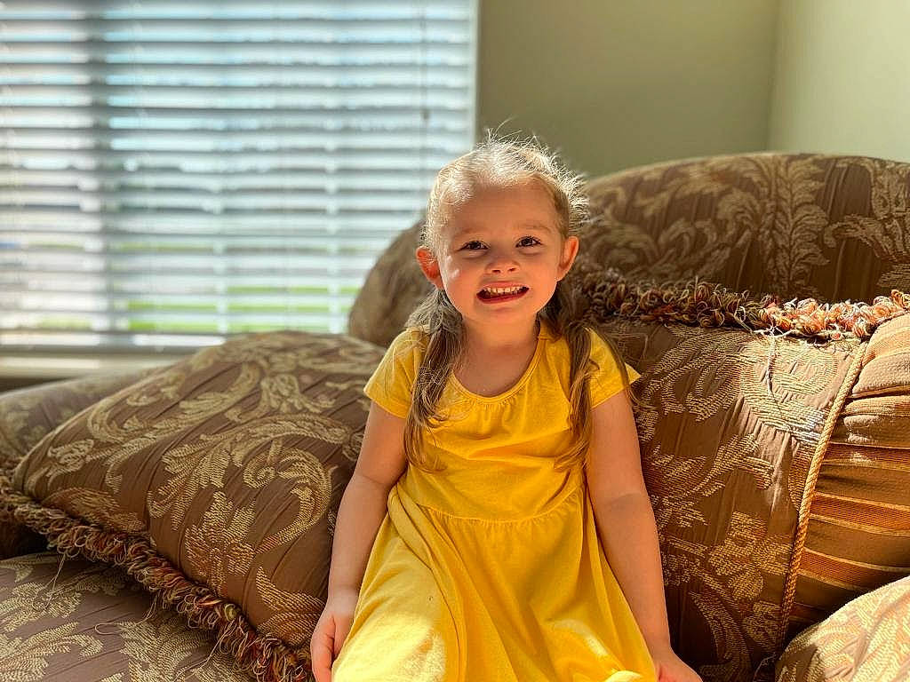 Emma joined the competition — help win amazing prizes! comfort, couch, dress, event, eye, flooring, fun, hairstyle, happy, joy, linens, living_room, person, pillow, smile, studio_couch, textile, throw_pillow, toddler, window