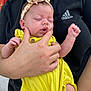 adidas_logo, adult_hand, baby, black_shirt, close_up, cute, face, fingers, floral_pattern, headband, holding, human, infant, outdoor, peaceful, person, portrait, skin, sleeping, yellow_outfit