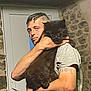 adult, animal, black_cat, cat, close_up, cozy, door, expression, furry, holding, home, indoor, man, person, pet, portrait, short_sleeve_shirt, stone_wall, tattoo, watch