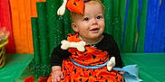 Josie joined the competition — help win amazing prizes! baby, bone_accessory, bow, celebration, child, colorful_background, costume, cute, decorations, festival, festive, happy, indoors, orange_dress, pink_socks, playful, portrait, rainbow, sitting, smiling