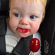 Josie is registered to the contest to win money with this photo: toddler, child, car_seat, lollipop, red_hair, blue_eyes, face, hand, candy, tongue, clothing, seatbelt, closeup, cute, infant, baby, person, indoor, expression, sitting