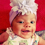 Oaklyann is registered to the contest to win money with this photo: baby, baby_girl, child, closeup, cute, expression, face, flower_headband, happy, headwear, indoors, infant, patterned_clothing, person, photography, portrait, red_background, skin, smiling, watermark