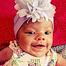 baby, baby_girl, child, closeup, cute, expression, face, flower_headband, happy, headwear, indoors, infant, patterned_clothing, person, photography, portrait, red_background, skin, smiling, watermark