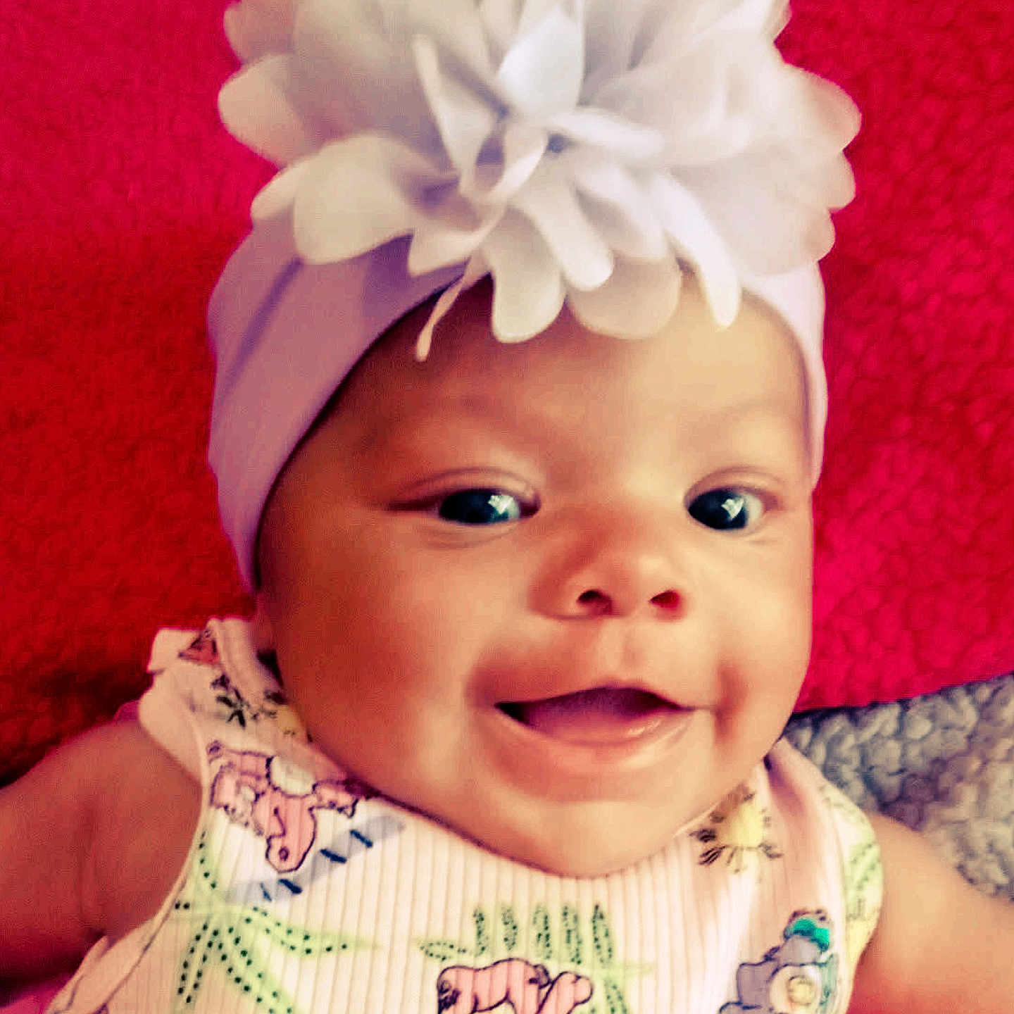 Oaklyann is registered to the contest to win money with this photo: baby, baby_girl, child, closeup, cute, expression, face, flower_headband, happy, headwear, indoors, infant, patterned_clothing, person, photography, portrait, red_background, skin, smiling, watermark
