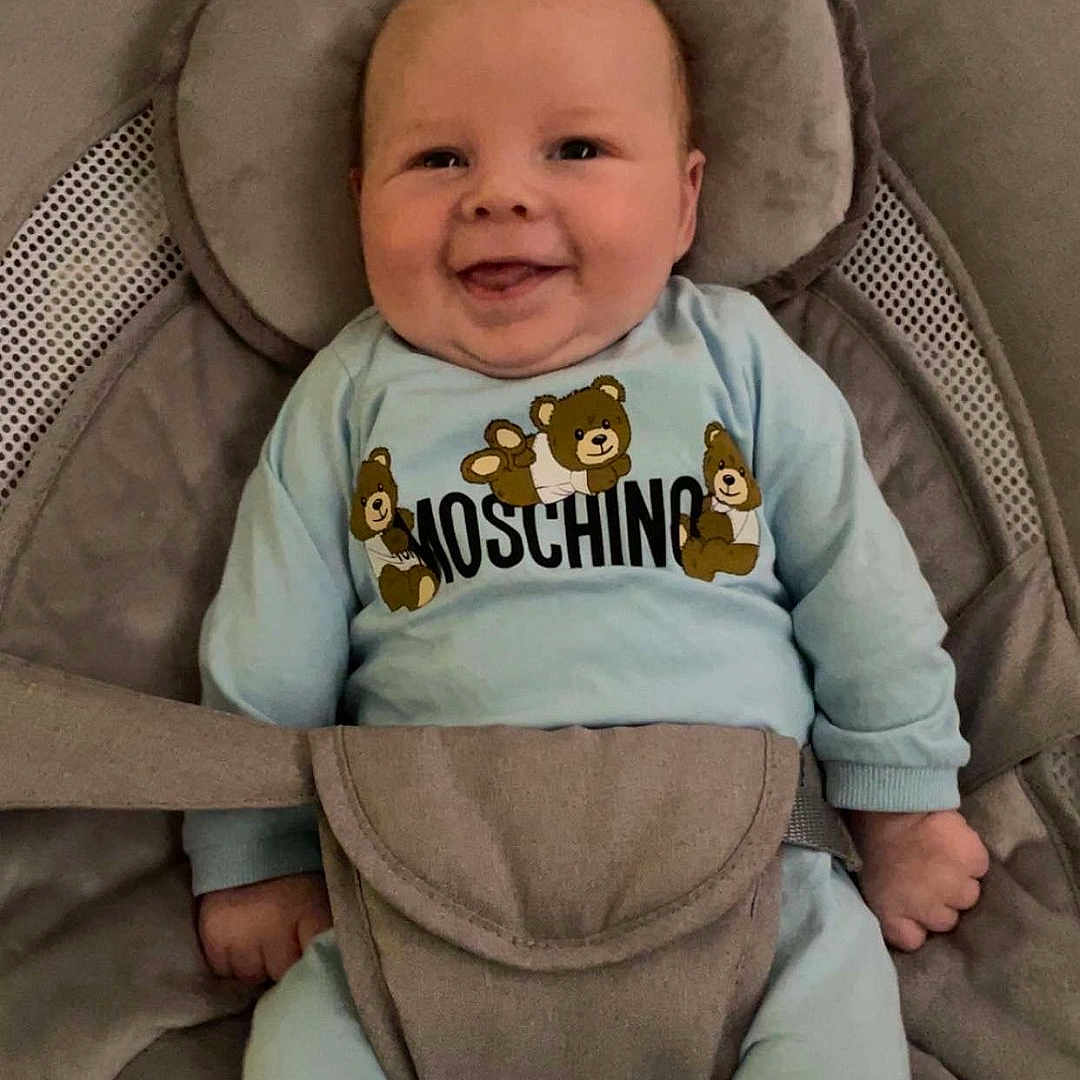 Tommy is registered to the contest to win money with this photo: baby, baby_seat, blue_clothing, child, comfort, cute, face, footwear, happy, head, indoor, infant, moschino, person, relaxed, seat, small_hands, smiling, straps, teddy_bear