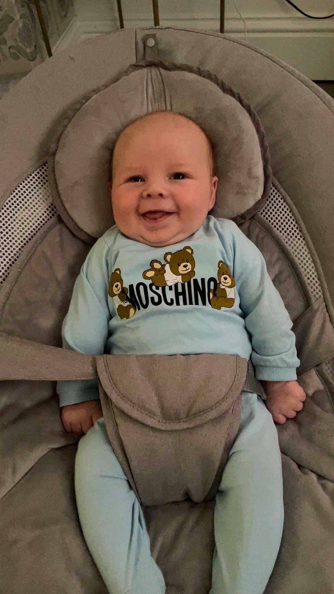 Tommy is registered to the contest to win money with this photo: baby, smiling, infant, blue_clothing, moschino, teddy_bear, baby_seat, straps, happy, cute, child, indoor, face, head, person, seat, comfort, relaxed, small_hands, footwear
