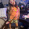 child, girl, sunglasses, peace_sign, orange_shirt, floral_shorts, slippers, living_room, couch, pillow, table, art_supplies, baby_car_seat, blanket, toy, carpet, indoor, playful, pose, smile