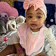 Beyon is registered to the contest to win money with this photo: baby, child, pink_bow, headband, bib, bracelet, plush_toys, bed, blanket, pillow, stuffed_animal, indoor, cute, smile, sitting, person, clothing, infant, toy, portrait