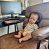 baby, headphones, chair, computer_monitor, keyboard, desk, water_dispenser, onesie, home_office, infant, footwearless, couch, indoor, technology, furniture, window, curtains, relaxed, person, expression