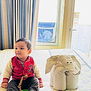 Aadvik joined the competition — help win amazing prizes! baby, barefoot, bed, bedspread, child, clothing, curtains, cute, elephant, furniture, home, indoor, portrait, red_jacket, sitting, smiling, toddler, towel_animal, toy, window