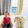 toddler, child, bed, towel_animal, elephant, curtains, window, indoor, smiling, sitting, clothing, red_jacket, barefoot, toy, bedspread, home, portrait, cute, baby, furniture