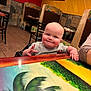 Blaine is registered to the contest to win money with this photo: baby, child, smiling, high_chair, restaurant, table, painted_table, colorful, indoor, furniture, person, cute, happy, face, skin, clothing, hand, seat, wood, booth