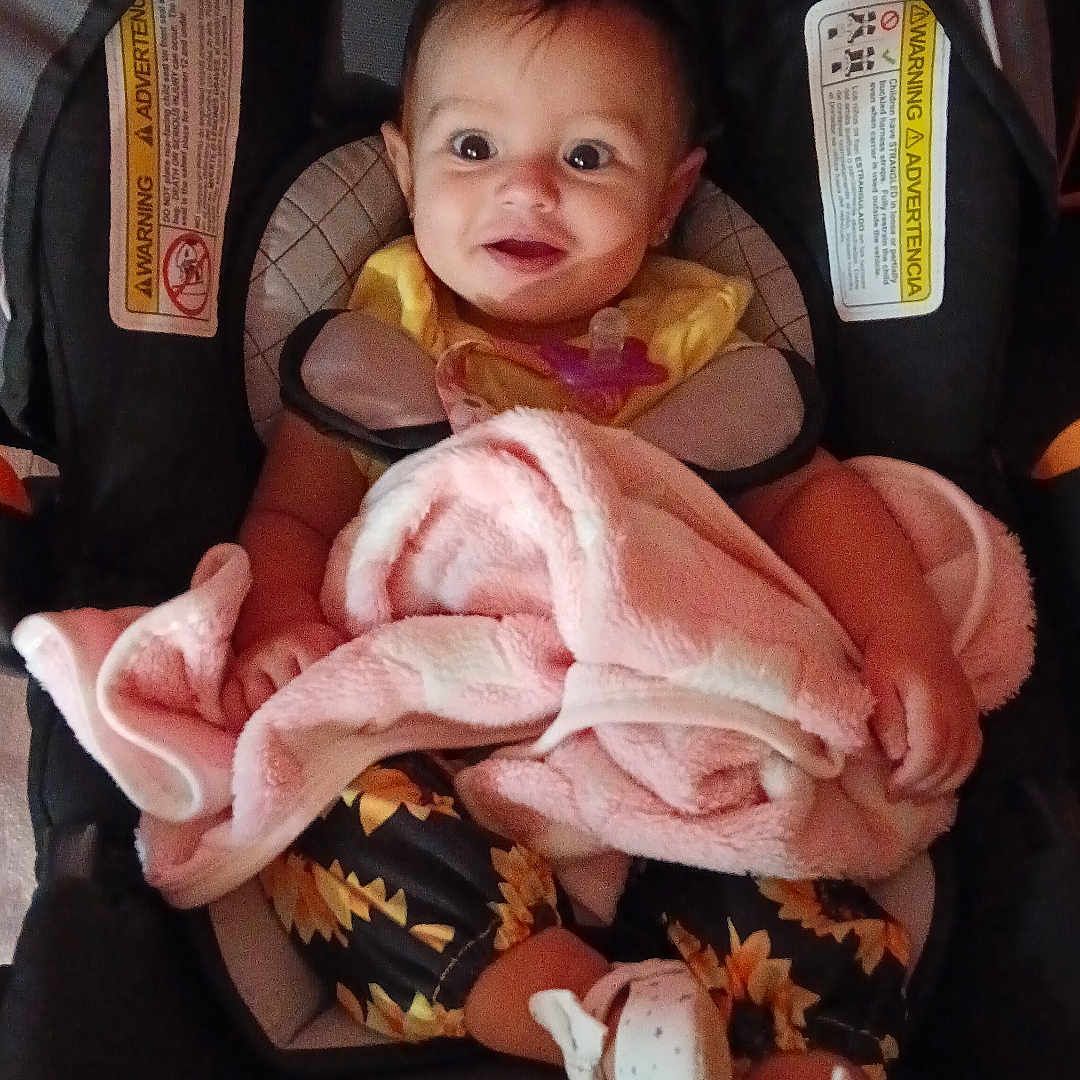 Ruby is registered to the contest to win money with this photo: baby, car_seat, blanket, infant, child, clothing, footwear, happy, face, person, sitting, indoors, yellow, pink, patterned, smile, cute, young, seatbelt, comfort
