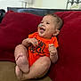baby, infant, smiling, orange_onesie, couch, red_cushion, happy, sitting, indoors, child, cute, face, feet, hand, expression, young, person, comfortable, playful, joyful