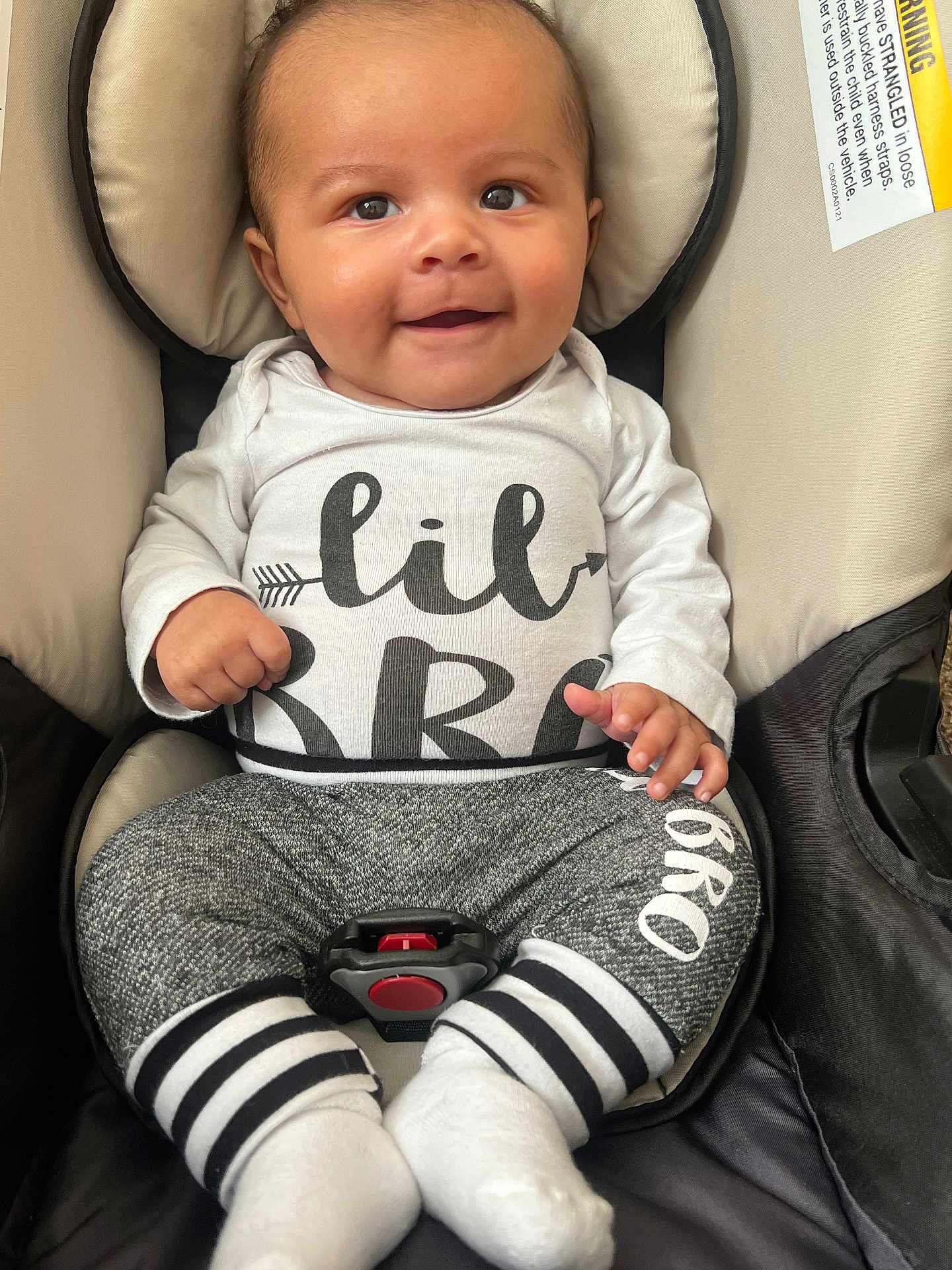 Malakai is registered to the contest to win money with this photo: baby, smiling, infant, car_seat, clothing, white_shirt, gray_pants, socks, baby_boy, happy, cute, child, face, portrait, seated, indoor, baby_fashion, baby_accessory, car_safety, closeup