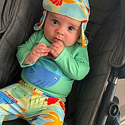 Romeo is registered to the contest to win money with this photo: baby, child, stroller, hat, colorful_clothing, fish_print, palm_tree_print, infant, cute, indoors, seated, looking_at_camera, hand_near_mouth, foot, headwear, fabric, patterned_clothes, person, casual_wear, soft_lighting
