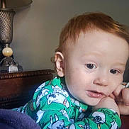 Kody is registered to the contest to win money with this photo: child, toddler, baby, portrait, indoors, sitting, blanket, pajamas, green_clothing, cartoon_print, red_hair, blue_eyes, face, hand, curious, serious_expression, home, lamp, furniture, cozy