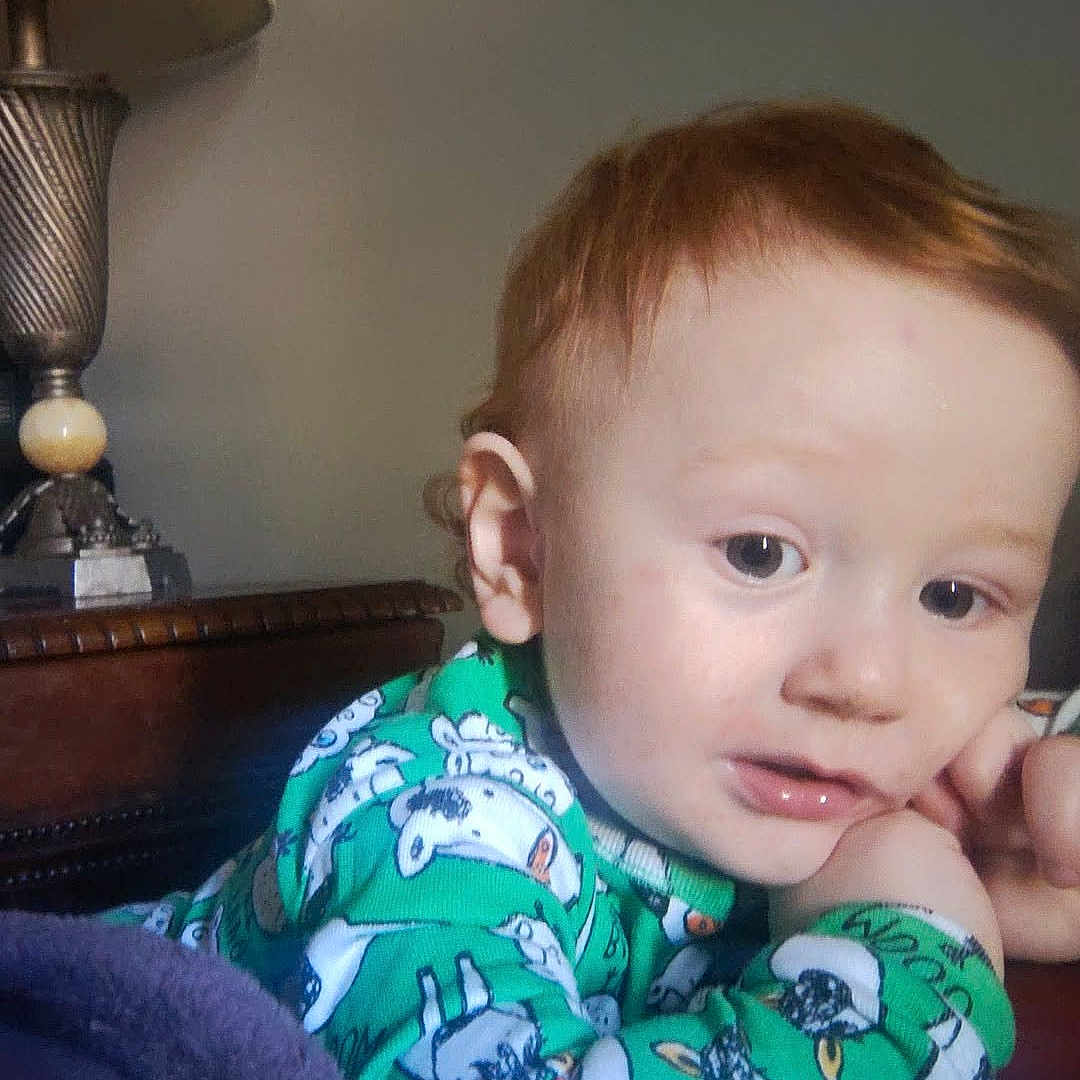 Kody is registered to the contest to win money with this photo: child, toddler, baby, portrait, indoors, sitting, blanket, pajamas, green_clothing, cartoon_print, red_hair, blue_eyes, face, hand, curious, serious_expression, home, lamp, furniture, cozy