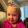 child, toddler, portrait, smiling, pink_shirt, hair_bow, blonde_hair, blue_eyes, rosy_cheeks, sitting, couch, cushion, indoors, home, warm_lighting, close_up, face, baby, happy, photograph