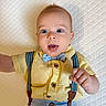 baby, child, infant, portrait, lying_down, indoors, mattress, quilted, yellow_shirt, bow_tie, suspenders, blue_pants, brown_buttons, wide_eyes, open_mouth, smiling, hand, arm, fashion, close_up