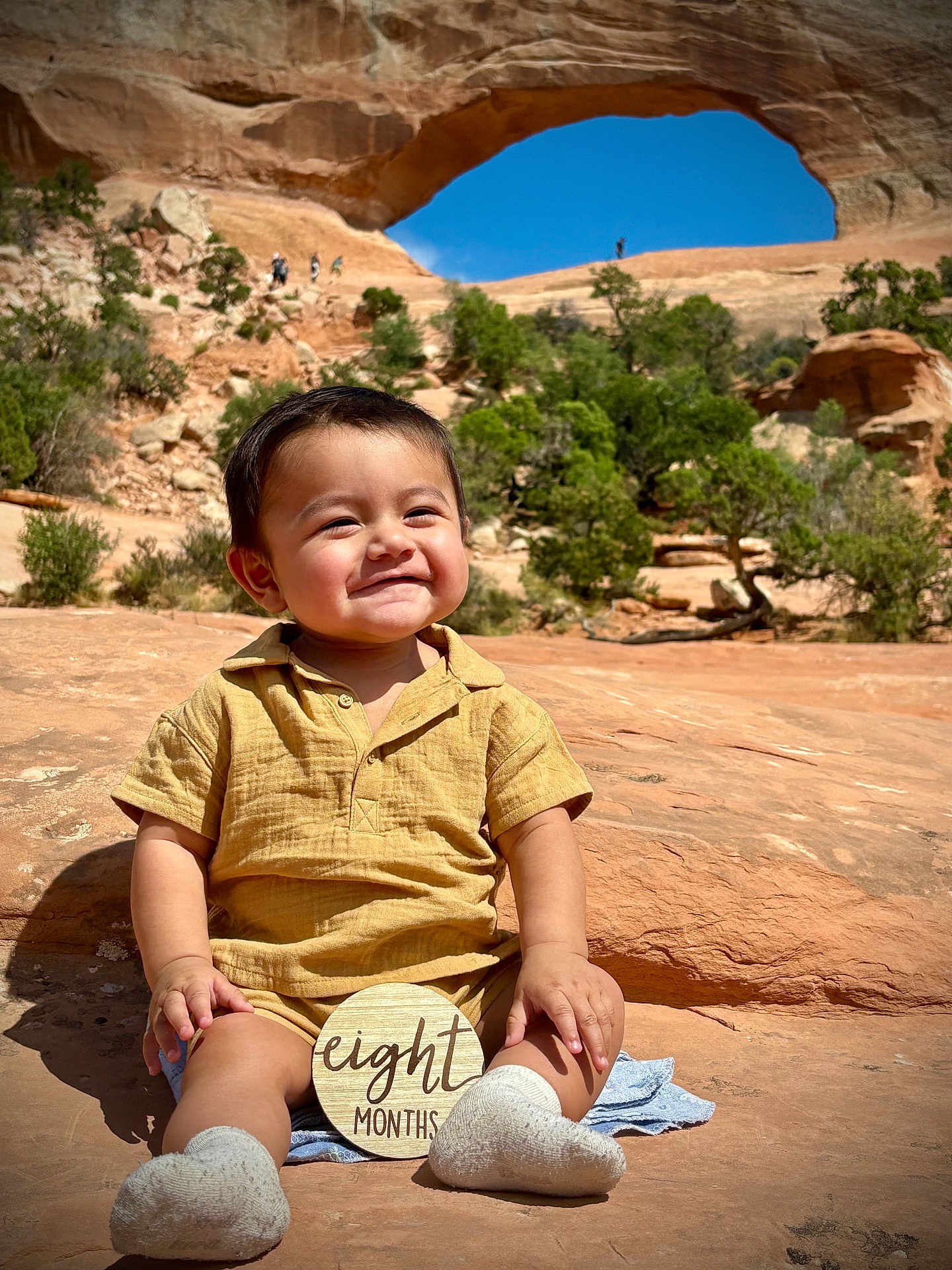 Joel is registered to the contest to win money with this photo: baby, child, outdoor, rock, natural_arch, sky, smile, milestone, sign, clothing, socks, nature, greenery, sunlight, portrait, hiking, desert, landscape, person, happy