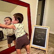 Joel joined the competition — help win amazing prizes! baby, child, mirror, reflection, floor, chalkboard, milestone, wooden_floor, wooden_frame, clothing, shorts, tank_top, indoor, standing, barefoot, smiling, home, room, door, sign