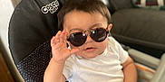 Joel is registered to the contest to win money with this photo: baby, child, sunglasses, high_chair, white_shirt, gray_pants, hand_gesture, indoor, living_room, couch, person, cute, fashion, seated, young_child, furniture, cosco_kids, portrait, funny, casual
