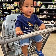 Asher joined the competition — help win amazing prizes! toddler, child, shopping_cart, blue_shirt, blue_sandals, grocery_store, produce, vegetables, shelves, floor, person, cute, indoor, holding_cart, shorts, expression, face, hair, seat, casual_clothing