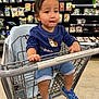 toddler, child, shopping_cart, blue_shirt, blue_sandals, grocery_store, produce, vegetables, shelves, floor, person, cute, indoor, holding_cart, shorts, expression, face, hair, seat, casual_clothing