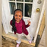 beige_pants, braided_hair, casual_clothing, child, daylight, door, frilly_socks, happy, home, maroon_top, outdoor, peace_sign, person, pink_sneakers, playful, porch, smiling, standing, wooden_floor, young_child